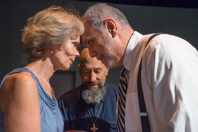 Juliet and Romeo lean their foreheads against each other, staring in each other's eyes, she in lose blue dress, he in white shirt, tie and suspenders, a bearded Lawrence with a blue shirt and gold cross around his neck looks down at his marriage book.